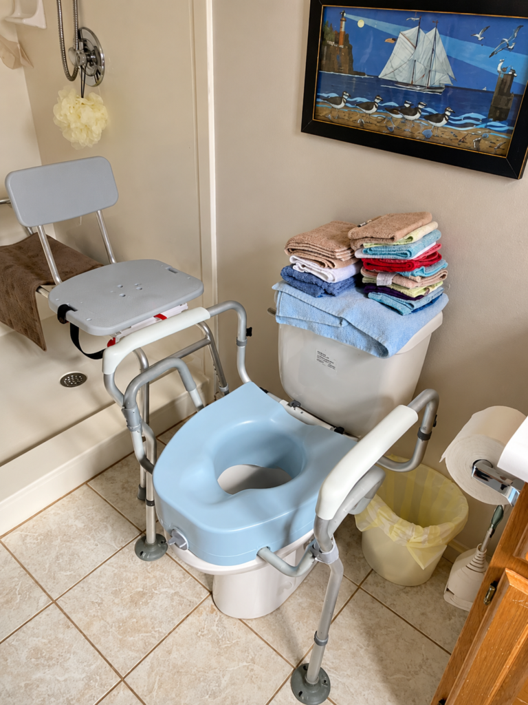 Accessible bathroom featuring a raised toilet seat with safety armrests, a shower chair, and other bathroom safety aids for seniors.