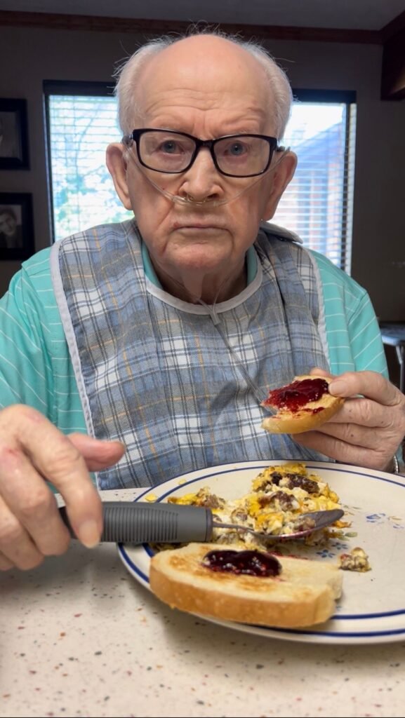 My dad eats breakfast at the kitchen table using a large-grip adaptive utensil. He is wearing glasses, a bib, and a nasal oxygen cannula while holding a piece of toast with jelly, showing how adaptive eating aids can help maintain independence.