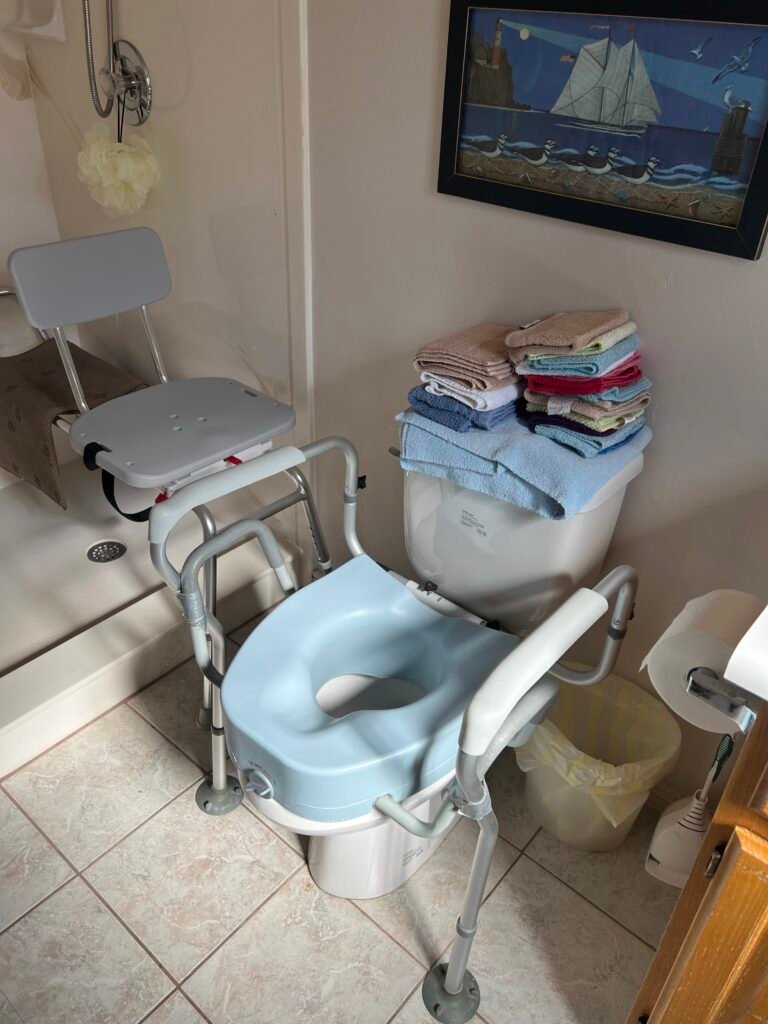 Dad's bathroom featuring a raised toilet seat with padded armrests and a toilet safety rail beside a walk-in bathtub with a shower chair. These bathroom safety aids helped him use the toilet more safely and maintain his independence at home.