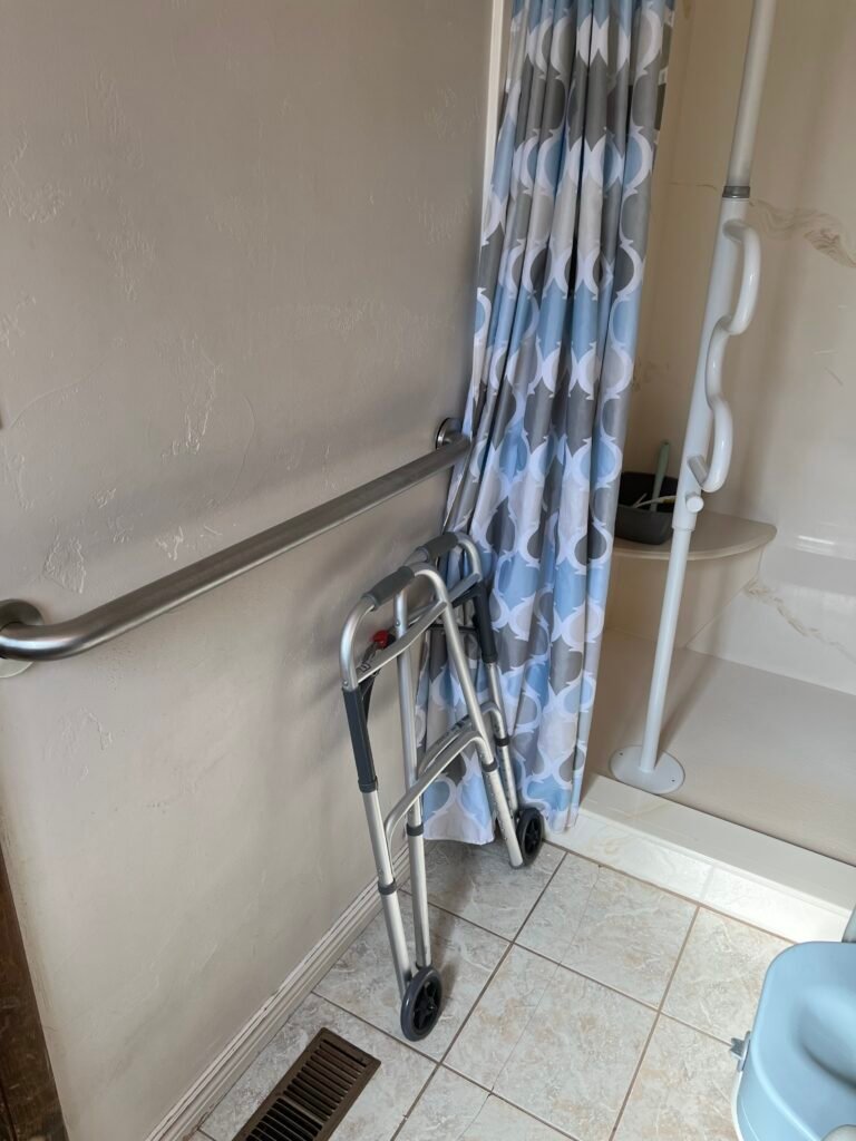 Dad's bathroom with a wall-mounted grab bar, a folding walker, and a floor-to-ceiling transfer pole beside the walk-in shower. These mobility and bathroom safety aids helped him stand, transfer, and move around more safely and independently.
