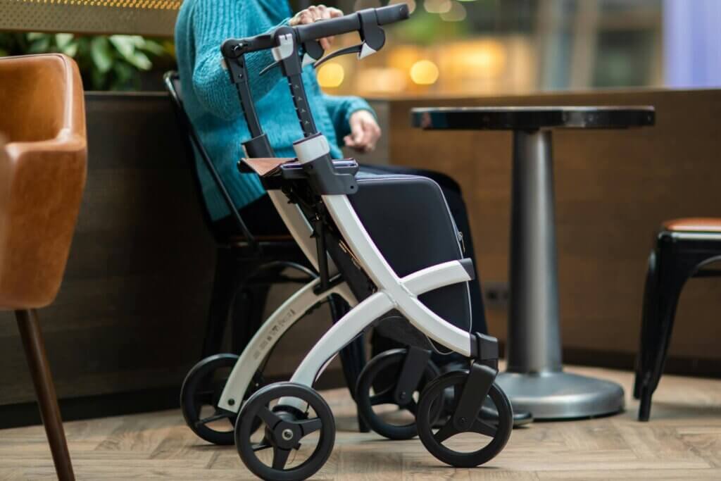 White Rollator walker with shopper bag next to a woman sitting at a table