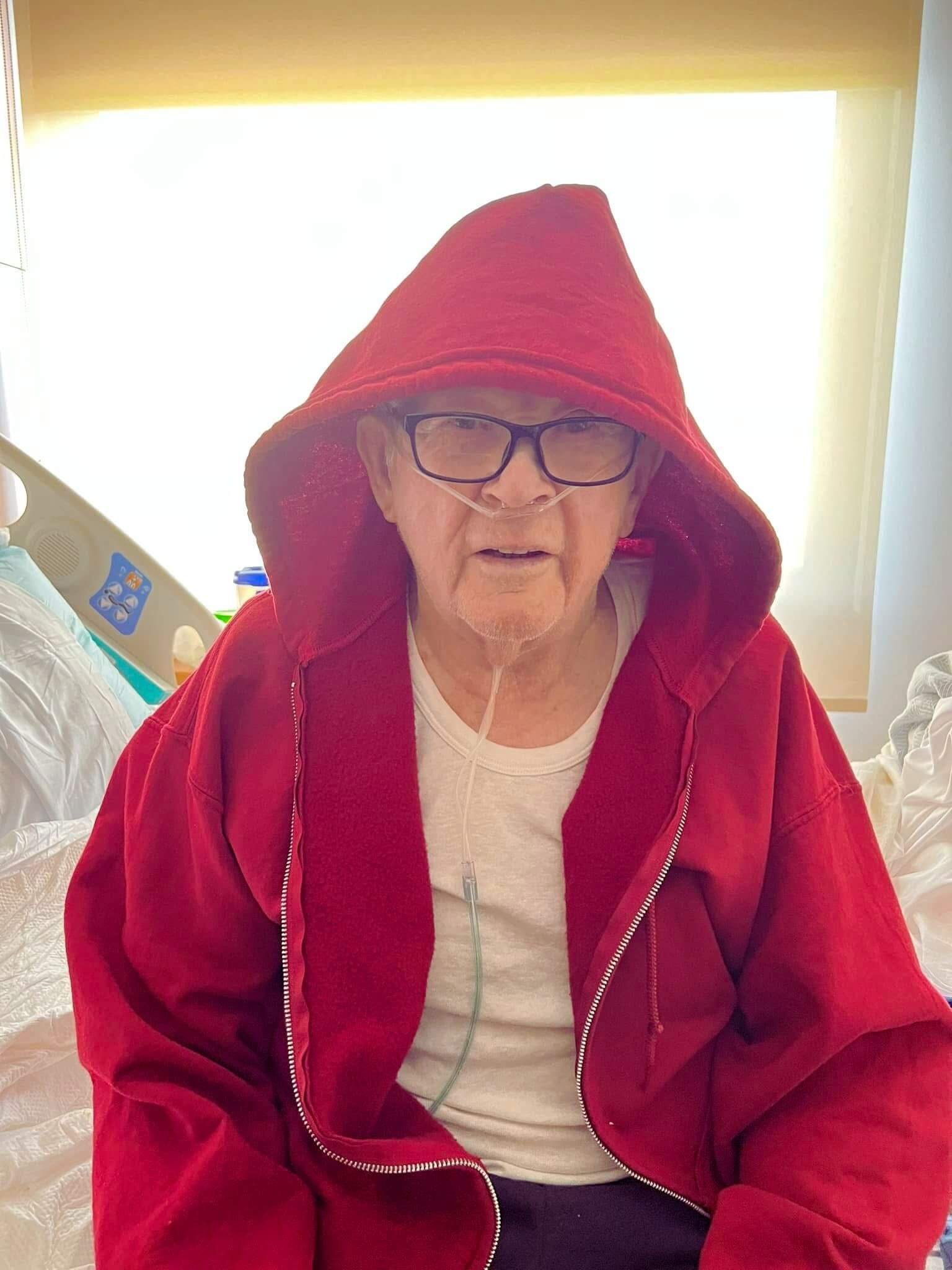 Ron's father sitting in a hospital room wearing a red hooded sweatshirt and receiving oxygen through a nasal cannula while preparing to be discharged home.