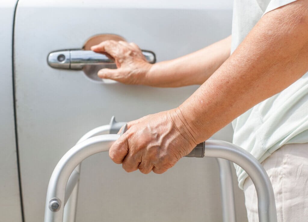 Older adult using a walker while reaching for a vehicle door handle.