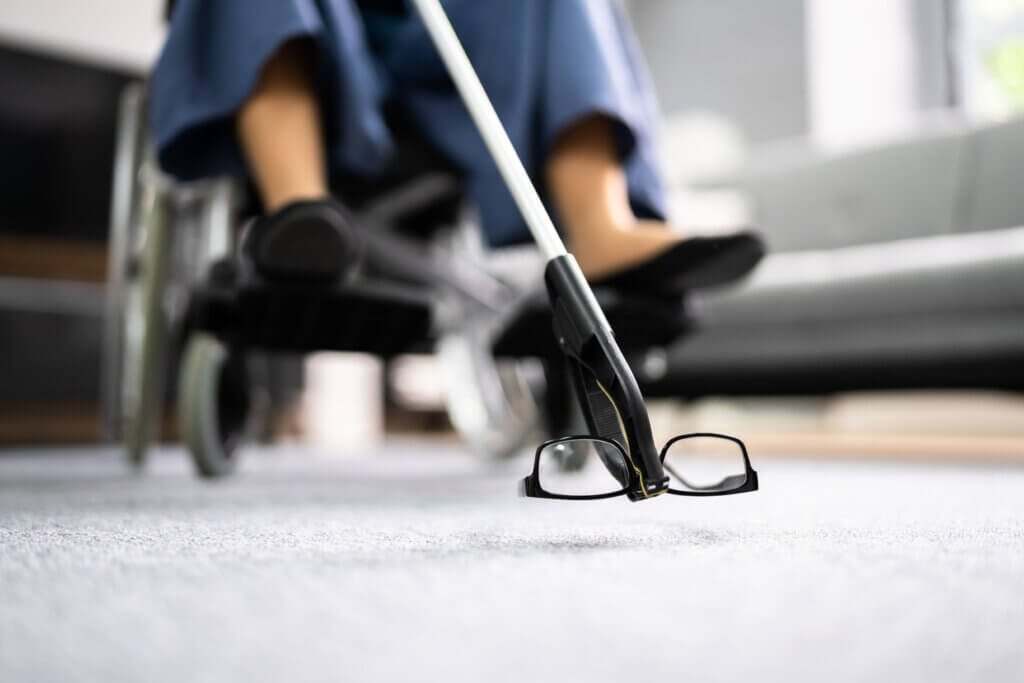 Wheelchair user using a reacher grabber tool to pick up eyeglasses from the floor without bending or reaching.