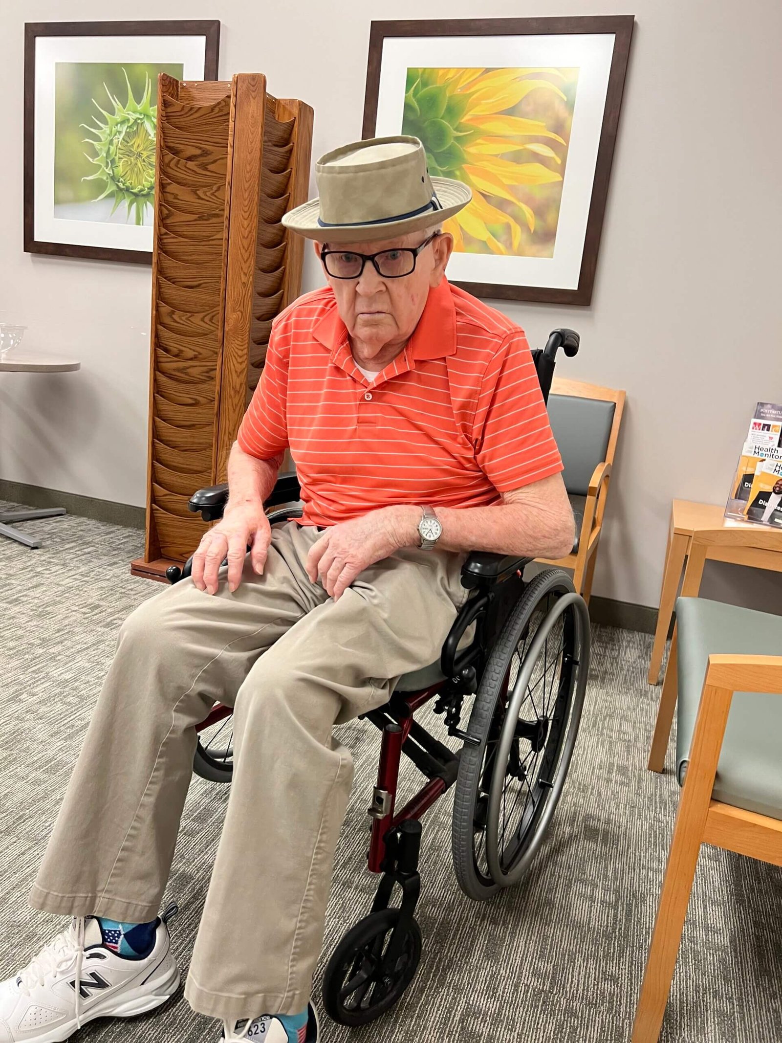 Ron's father sitting in his wheelchair while waiting for a doctor's appointment, wearing a sun hat and glasses in a medical office waiting room.