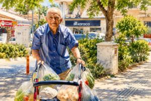 An older man pushes a red rollator loaded with grocery bags while walking along a sunny neighborhood sidewalk, demonstrating how mobility aids support independent shopping and daily living.